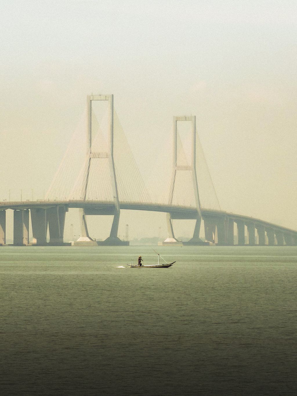 The cable-stayed Suramadu Bridge in pale haze over green water, a lone fisherman standing in a small wooden boat beneath its two towers.