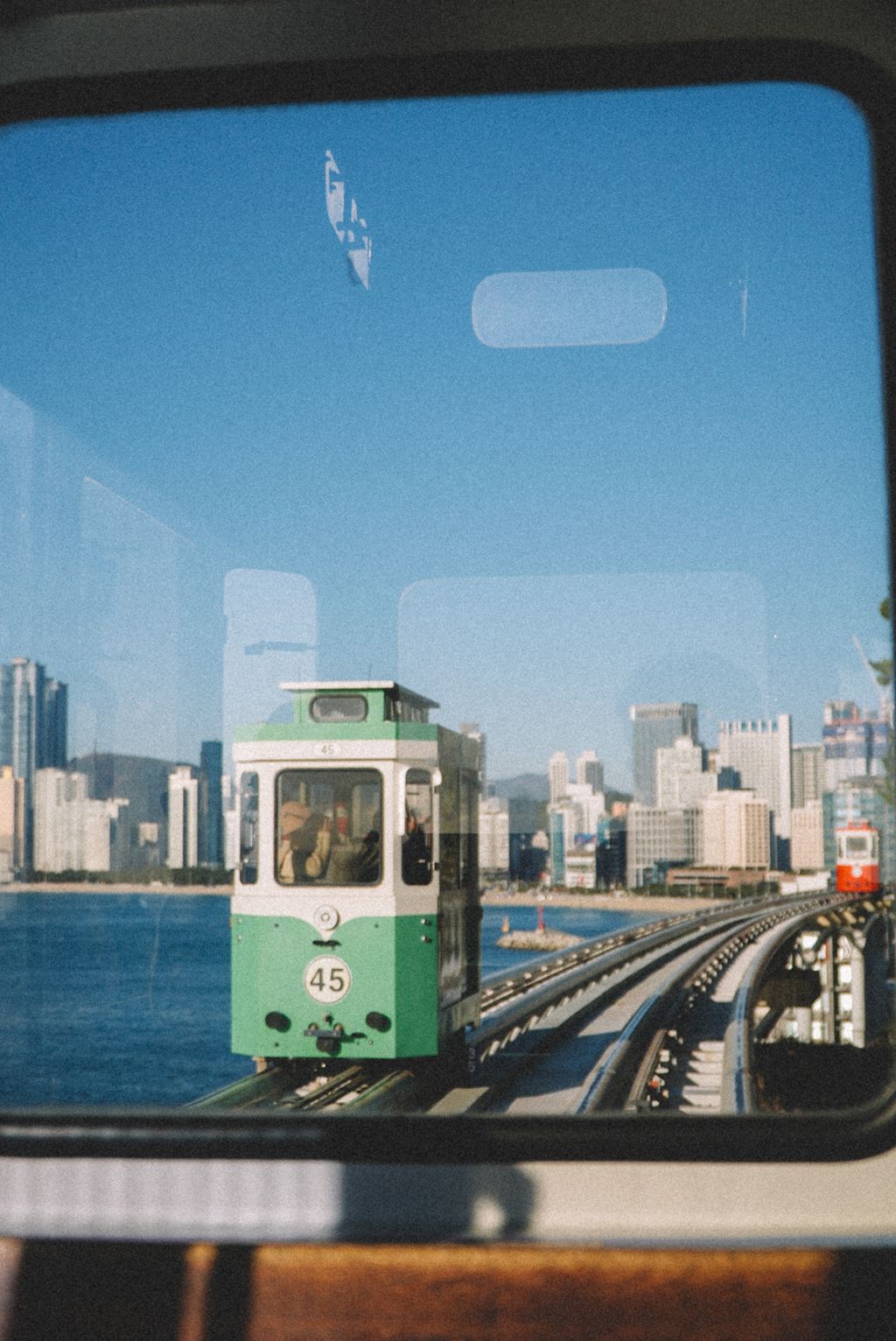 A green vintage-style beach train, number 45, approaching on elevated coastal tracks, seen through a train window with Busan's high-rises and the sea behind.