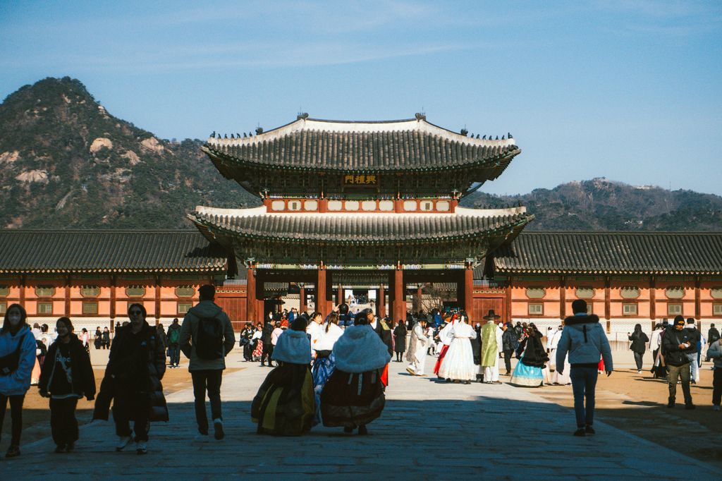 Visitors in hanbok and winter coats walking toward Heungnyemun Gate at Gyeongbokgung Palace, mountains behind under a clear blue sky.