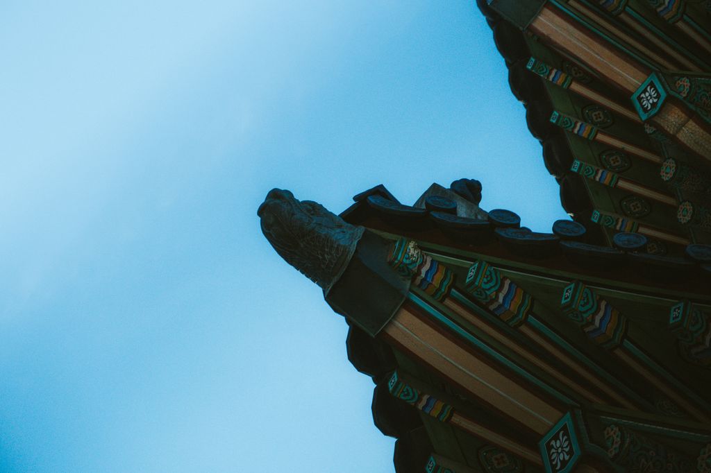 Low-angle detail of a palace roof corner: a carved stone guardian at the tip of the eave and painted rafters against a pale blue sky.